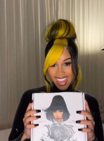 Cardi B poses with an album. Her hair is yellow and black and she wears it tied up in a high bun.